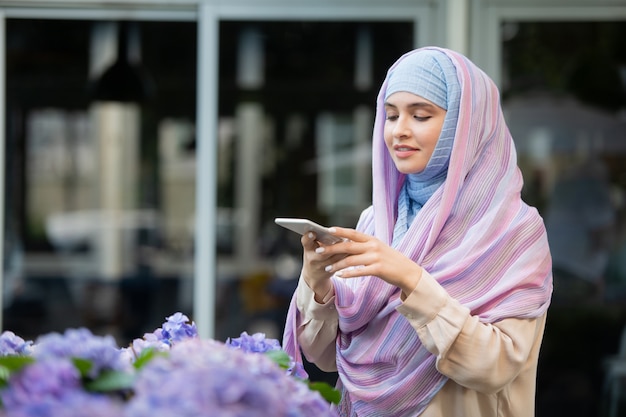 Joven mujer móvil en hijab tomando fotos de flores en el teléfono inteligente mientras pasa tiempo al aire libre