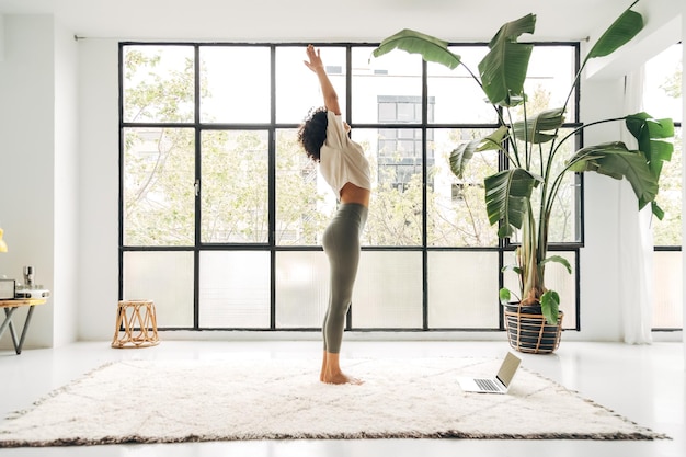 Joven mujer multirracial haciendo el saludo al sol en casa con un video tutorial en Internet usando una computadora portátil Práctica de yoga Surya namaskar Copiar espacio