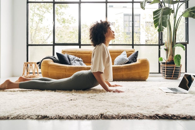 Joven mujer multirracial haciendo yoga en casa siguiendo la lección de tutorial en línea Bhujangasana cobra pose