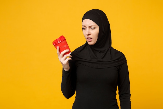 Joven mujer musulmana árabe molesta triste cansada en ropa negra hijab sostenga una taza de café de papel aislada en el retrato de pared amarilla. Concepto de estilo de vida religioso de la gente.