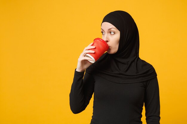 Joven mujer musulmana árabe molesta triste cansada en ropa negra hijab sostenga una taza de café de papel aislada en el retrato de pared amarilla. Concepto de estilo de vida religioso de la gente.