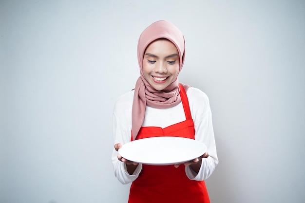 Joven mujer musulmana asiática en delantal de pie y sosteniendo un plato blanco vacío