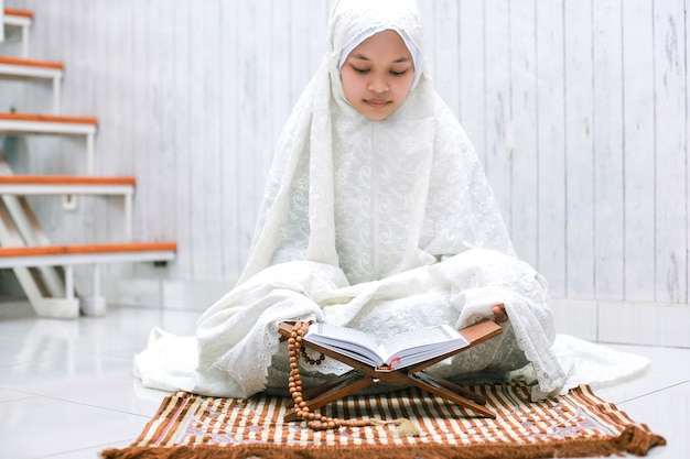 Joven mujer musulmana asiática leyendo el libro sagrado AlQuran en la estera de oración en casa