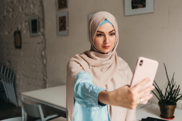 Joven mujer musulmana bastante moderna en hijab trabajando en la computadora portátil en la sala de oficina, educación en línea