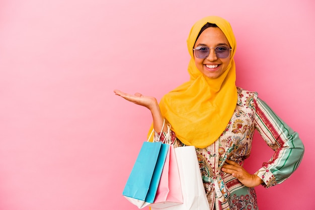 Joven mujer musulmana comprando ropa aislada sobre fondo rosa mostrando un espacio de copia en una palma y sosteniendo otra mano en la cintura.