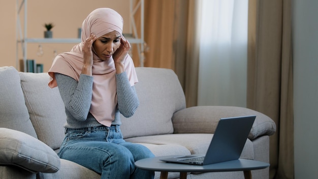 Joven mujer musulmana en hijab rosa sentada en un sofá trabajando usando una computadora portátil con dolor de cabeza migraña triste