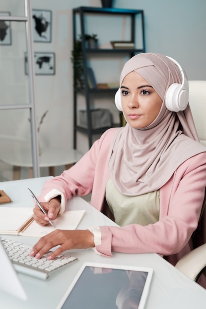 Joven mujer musulmana seria en auriculares, hijab y ropa casual elegante sentada en la mesa frente al monitor de la computadora y tomando notas