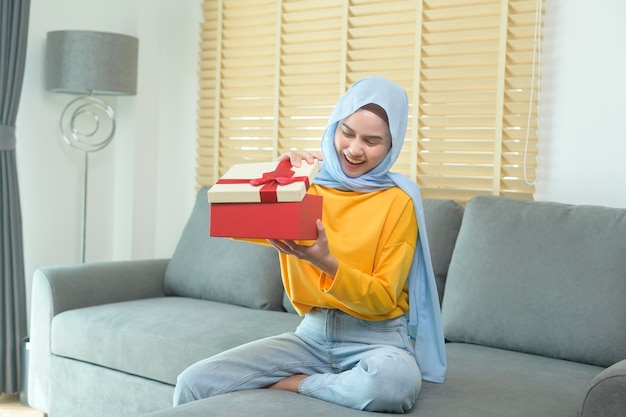 Joven mujer musulmana sorprendida abriendo una caja de regalo en la sala de estar