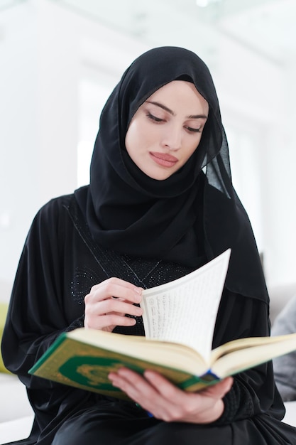 joven mujer musulmana tradicional leyendo el Corán en el sofá antes de la cena iftar durante una fiesta de ramadán en casa