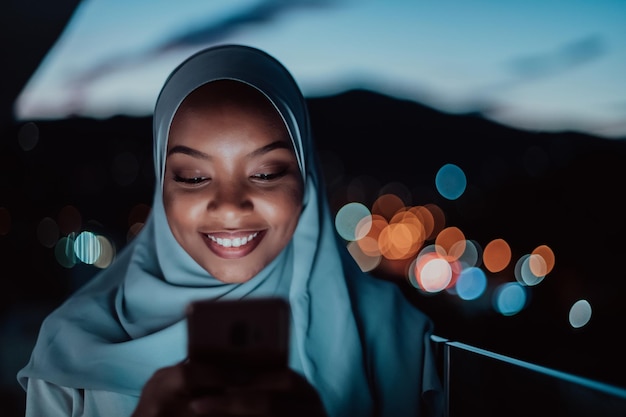 Joven mujer musulmana con velo de bufanda en la calle urbana de la ciudad por la noche enviando mensajes de texto en un teléfono inteligente con luz de la ciudad en el fondo. foto de alta calidad
