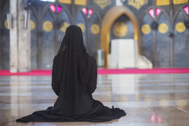 Joven mujer musulmana en vestido negro de desgaste sentado solo en la mezquita, vista trasera.