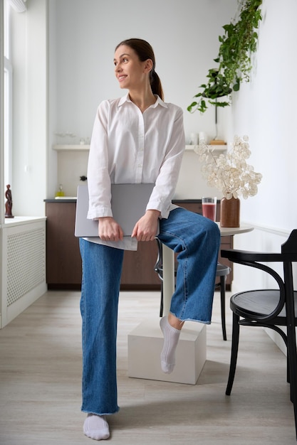 Joven mujer de negocios alegre y hermosa sonriendo con camisa blanca y jeans usando una computadora portátil en el interior