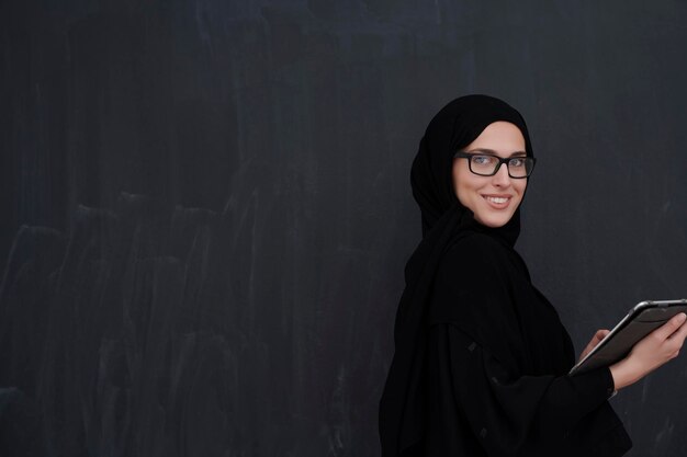 Joven mujer de negocios árabe con ropa tradicional o abaya y anteojos sosteniendo una tableta frente a una pizarra negra que representa la moda y la tecnología islámicas modernas