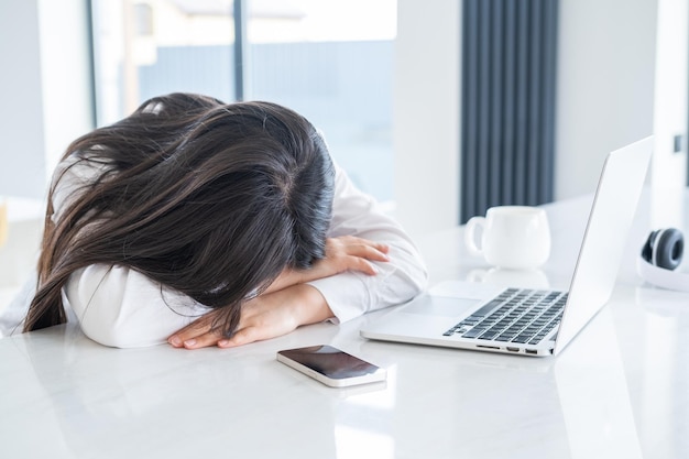 Joven mujer de negocios asiática cansada con cabello largo trabajando en una computadora portátil en una oficina en casa brillante