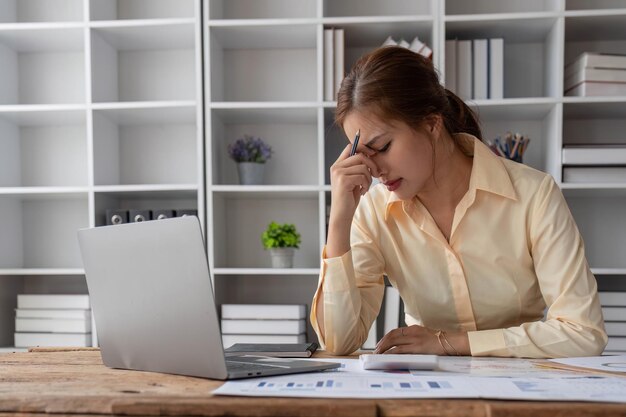 Joven mujer de negocios asiática que trabaja en una computadora portátil estresada tiene dolor de cabeza y piensa duro desde el trabajo en la oficina