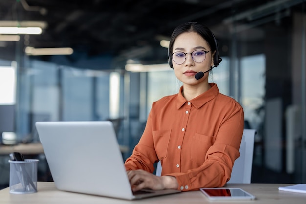 Joven mujer de negocios asiática seria usando portátil y auriculares en la oficina