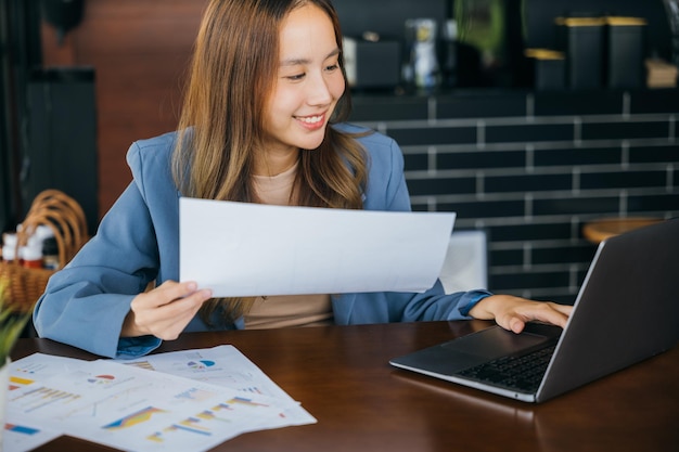 Una joven mujer de negocios asiática sonriente leyendo un documento de informe comparado con una computadora portátil Mujer contable feliz revisando datos en tablas y gráficos financieros afuera en una cafetería