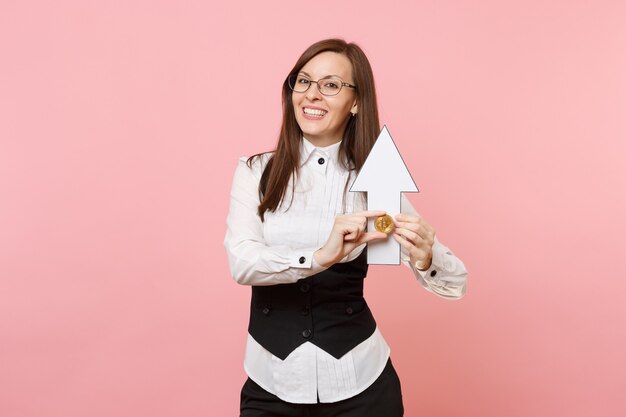 Joven mujer de negocios atractiva alegre en traje negro, gafas con bitcoin, flecha hacia arriba aislada sobre fondo rosa pastel. Jefa. Concepto de riqueza de carrera de logro. Copie el espacio para publicidad.