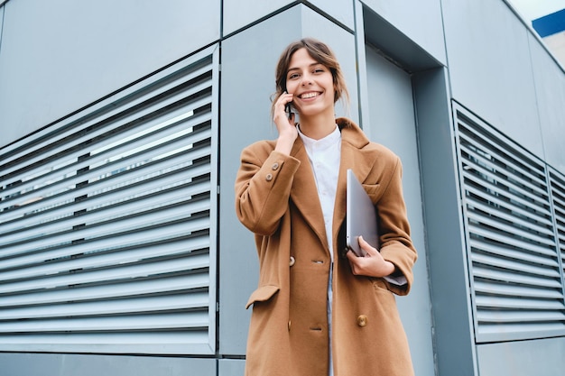 Joven mujer de negocios bonita con abrigo y portátil mirando alegremente a la cámara mientras habla por teléfono móvil al aire libre