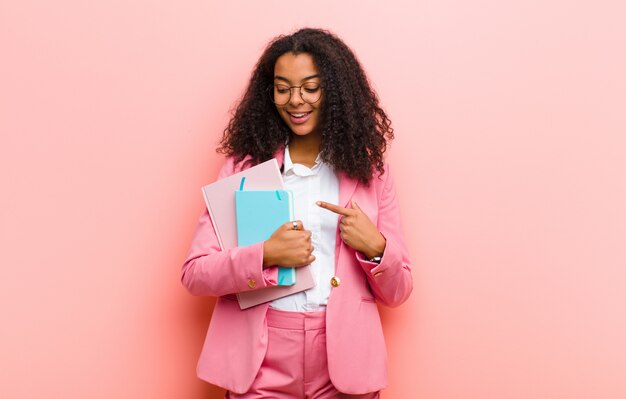 Joven mujer de negocios bonita negra con libros contra la pared rosa