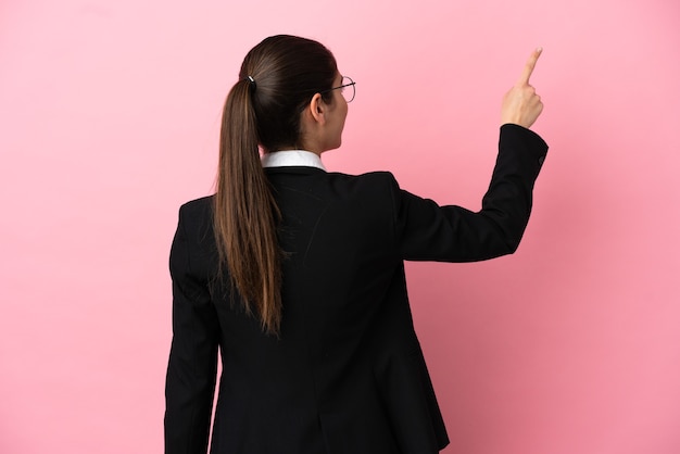 Joven mujer de negocios caucásica aislada sobre fondo rosa apuntando hacia atrás con el dedo índice