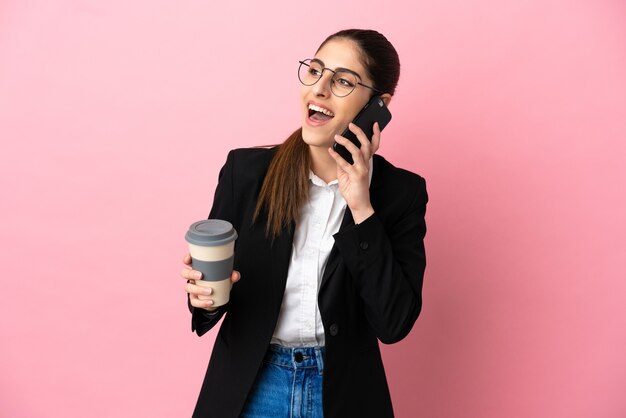 Joven mujer de negocios caucásica aislada sobre fondo rosa con café para llevar y un móvil