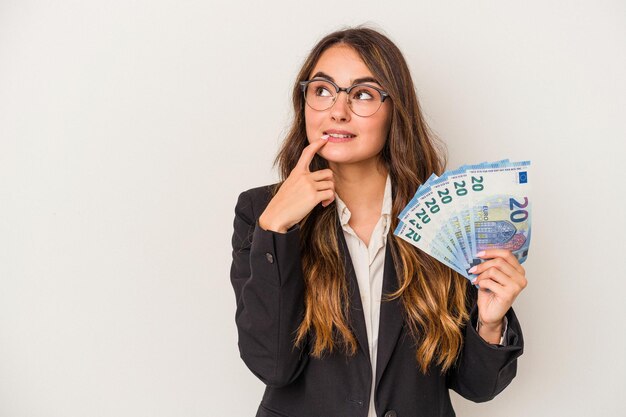 Joven mujer de negocios caucásica sosteniendo billetes aislados en fondo blanco relajado pensando en algo mirando un espacio de copia.