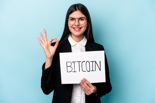 Joven mujer de negocios caucásica sosteniendo un cartel de bitcoin aislado sobre fondo azul alegre y confiada mostrando un gesto correcto