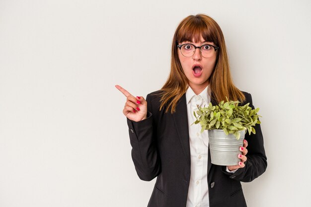 Joven mujer de negocios caucásica sosteniendo una planta aislada sobre fondo blanco apuntando hacia el lado