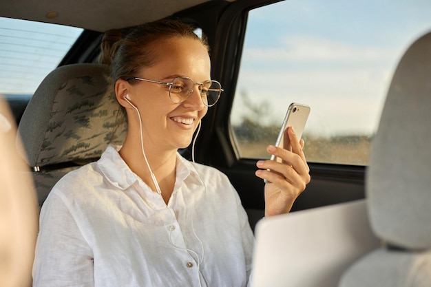 Joven mujer de negocios caucásica usando un teléfono inteligente y sonriendo mientras se sienta en el asiento trasero del auto navegando por Internet leyendo mensajes en las redes sociales