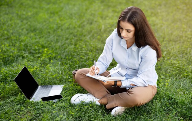 Joven mujer de negocios centrada sentada con las piernas cruzadas en la hierba con una computadora portátil a su lado