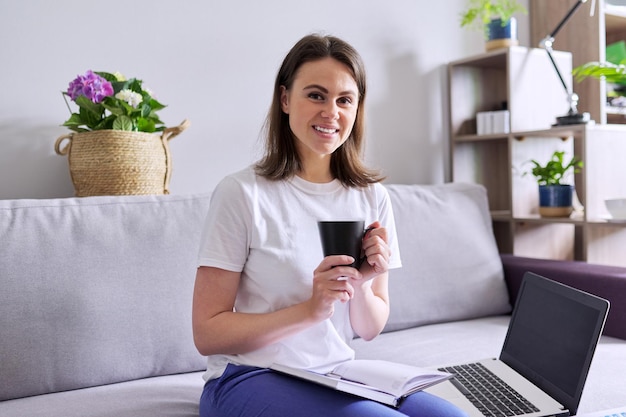 Joven mujer de negocios con computadora portátil, taza de café sentada en el sofá, fondo interior de la casa. Empleo remoto, autónomo, educación y enseñanza en línea, lecciones de carrera y concepto de desarrollo