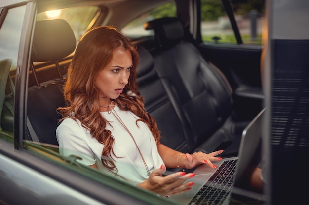 Joven mujer de negocios de confianza escribiendo en su computadora portátil en el asiento trasero del auto.