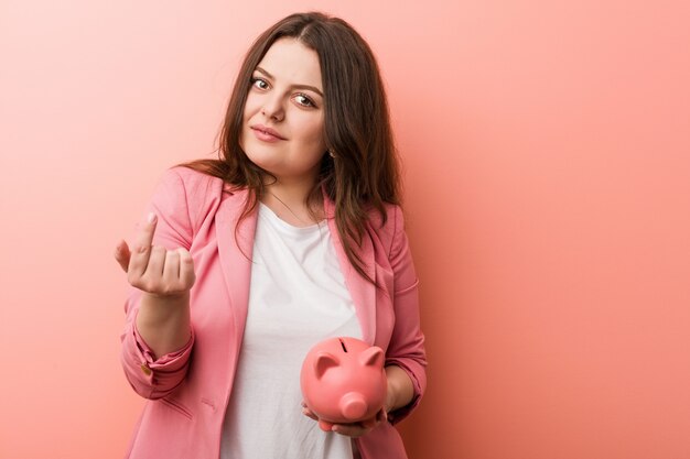 Foto joven mujer de negocios con curvas de talla grande sosteniendo una alcancía apuntando con el dedo hacia usted como si invitara a acercarse