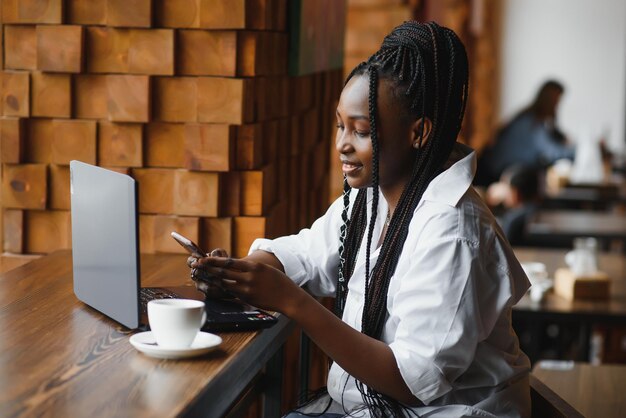Una joven mujer de negocios está organizando la próxima reunión de negocios en un moderno espacio de coworking usando una computadora portátil. Mujer joven independiente está leyendo correos electrónicos y otros mensajes en un teléfono móvil.