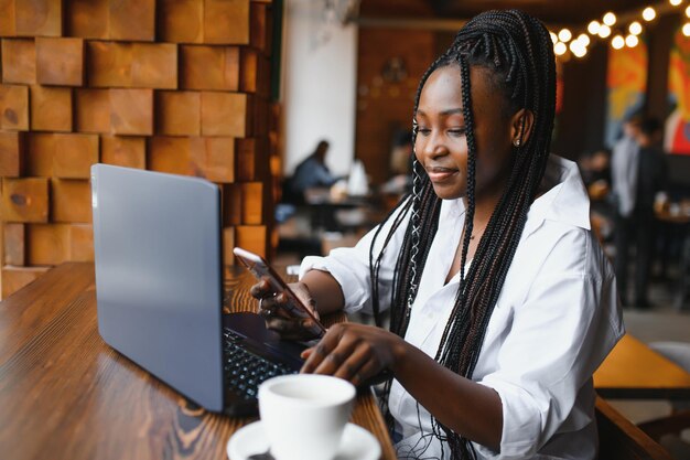 Una joven mujer de negocios está organizando la próxima reunión de negocios en un moderno espacio de coworking usando una computadora portátil. Mujer joven independiente está leyendo correos electrónicos y otros mensajes en un teléfono móvil.