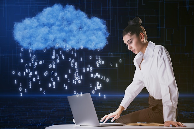 Joven mujer de negocios europea atractiva que usa una computadora portátil mientras está sentada en un escritorio con un holograma de nube creativa con código binario lluvia en un fondo azul borroso Concepto de computación y tecnología en la nube
