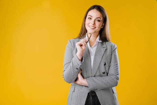 Una joven mujer de negocios feliz con chaqueta y camisa con gafas sonríe y posa en la cámara con fondo amarillo