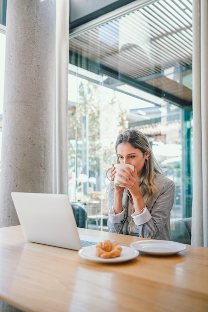 Joven mujer de negocios feliz haciendo videollamadas, bebiendo café de una taza en un lugar de trabajo moderno y abierto