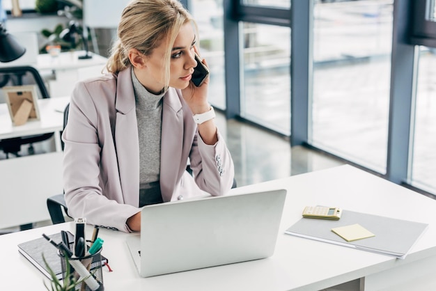 joven mujer de negocios hablando por teléfono inteligente y usando portátil en la oficina