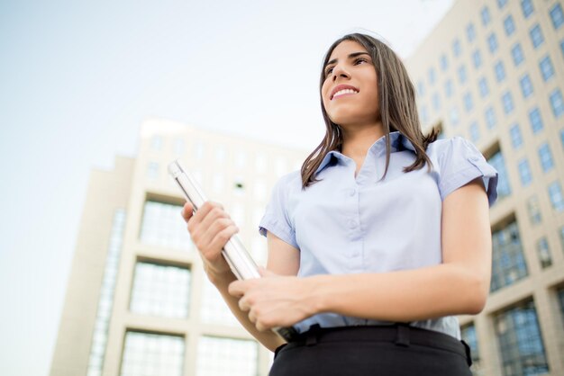 Joven mujer de negocios hermosa con una laptop en las manos, parada frente al edificio de oficinas y sonriendo mirando a lo lejos.