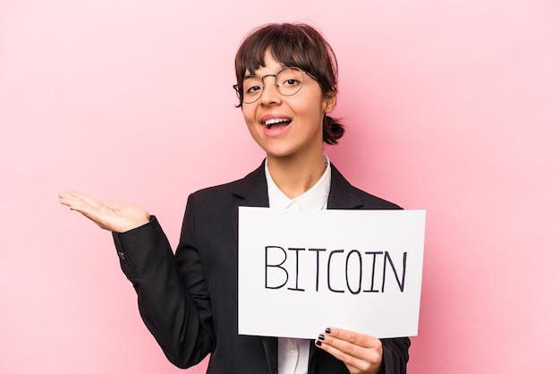 Joven mujer de negocios hispana sosteniendo un cartel de bitcoin aislado en un fondo rosa mostrando un espacio de copia en una palma y sosteniendo otra mano en la cintura
