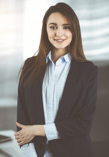 Una joven mujer de negocios inteligente con una chaqueta azul está parada en su lugar de trabajo. Retrato de un especialista en oficina soleada.