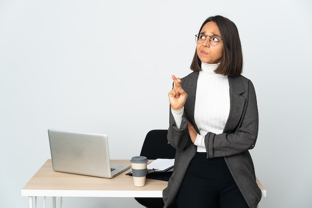 Joven mujer de negocios latina que trabaja en una oficina aislada sobre fondo blanco con los dedos cruzando y deseando lo mejor