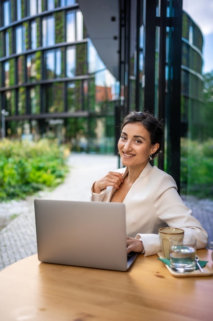 Joven mujer de negocios morena sonriente con elegante vestido inteligente trabajando en una laptop en un café en la calle de