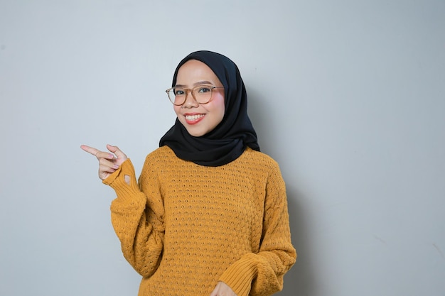 joven mujer de negocios musulmana asiática con suéter naranja y gafas que señala el espacio en blanco a un lado