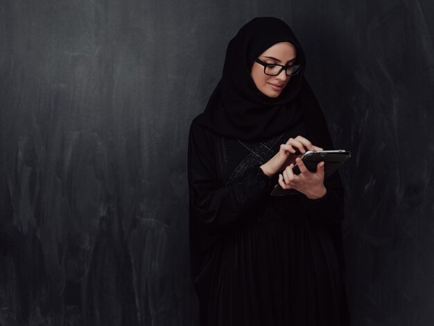 Joven mujer de negocios musulmana moderna que usa un teléfono inteligente con ropa hijab frente a una pizarra negra.