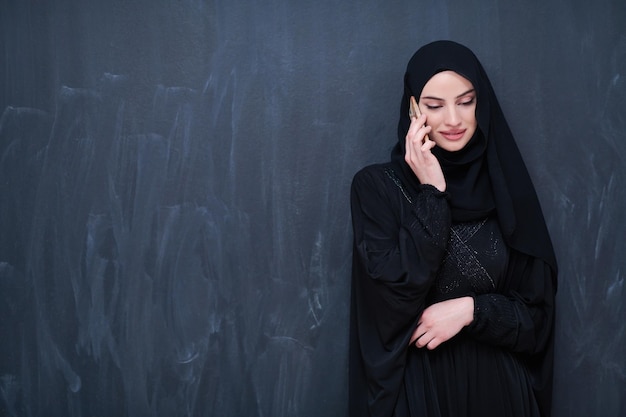 joven mujer de negocios musulmana moderna que usa un teléfono inteligente con ropa hiyab frente a una pizarra negra