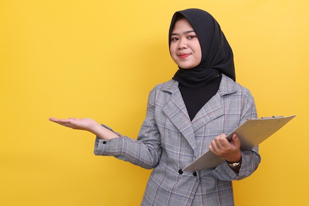 Una joven mujer de negocios musulmana que lleva pañuelo en la cabeza muestra un gesto de saludo y bienvenida mientras sostiene c
