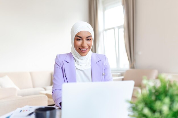 Joven mujer de negocios musulmana con ropa informal elegante discutiendo negocios y sonriendo mientras está sentada en el coworking creativo